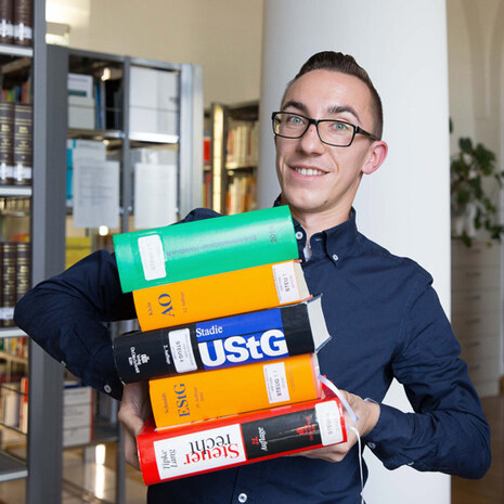 Auf dem Bild ist ein junger Kollege abgebildet, welcher in einem Finanzamt mit einer Vielzahl von übereinander gestapelten Gesetzesbüchern (AO, UStG, EStG) vor der Kamera steht. Das Bild soll vermitteln, dass viele Gesetze das Steuerrecht prägen aber denn