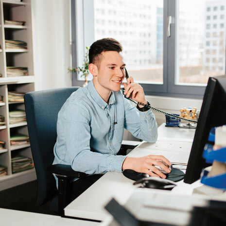 Auf dem Bild ist ein junger Kollege abgebildet, welcher an seinem Büroarbeitsplatz im Finanzamt sitzt. Er hält die Maus und den Telefonhörer in der Hand und schaut mit einem leichten Grinsen auf seinen Monitor. Das Bild steht für die Freude, welche der ju